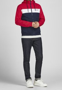 Rot und marinefarben blockierte Jacke mit Kapuze, Reißverschluss vorne und Logo, kombiniert mit dunklen Jeans und weißen Sneakers.