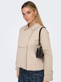 Vrouw met recht blond haar, een beige jas, een blauwe rok met bloemenpatroon, zilveren oorbellen en een kleine zwarte schoudertas.