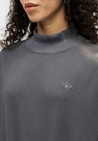 Γκρι φούτερ με ψηλό λαιμό και κεντημένο λογότυπο σε πιο ανοιχτό γκρι. Απαλή υφή και μινιμαλιστικός σχεδιασμός, κοντινή θέα στο επάνω μέρος του σώματος.