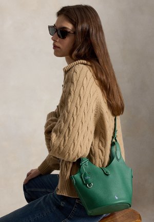 Femme aux longs cheveux bruns portant des lunettes de soleil et un pull en maille torsadée beige, assise et tenant un sac à main en cuir vert sur l'épaule.
