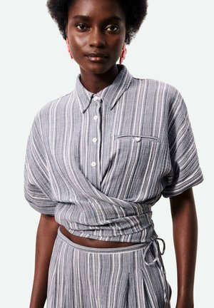 Vrouw draagt een grijs-wit gestreept wikkelshirt met korte mouwen en een bijpassende hoge taille rok, staand tegen een effen achtergrond.