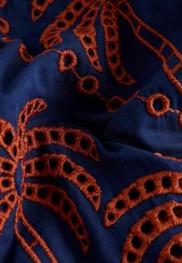 Tissu bleu profond avec une broderie orange rouille en relief formant des motifs circulaires et linéaires, montrant des plis et des détails de texture.