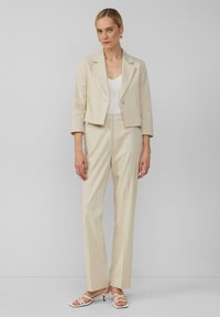 Blazer beige cintré avec une texture rayée subtile, associé à un pantalon beige clair à jambes droites et des talons blancs à lanières.