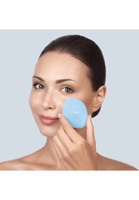 Outil facial bleu clair avec une forme lisse et arrondie, tenu contre un visage de teinte de peau, affichant le nom de la marque « GESKE » en texte blanc.