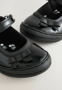 Chaussures noires en cuir verni avec un détail en nœud, bout rond et sangle ajustable. Semelle en caoutchouc texturée pour une meilleure adhérence. Doublure intérieure noire.