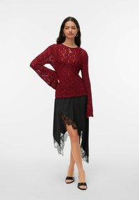 Bordowa koronkowa bluzka z długim rękawem w połączeniu z czarną asymetryczną spódnicą z koronkowym wykończeniem. Stylizację uzupełniają czarne klapki.