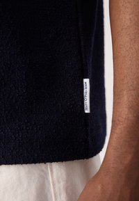 Marineblå, kort genser i myk terry-stoff med en teksturert overflate. Har et lite hvitt merke på siden med "Marc O’Polo Denim."