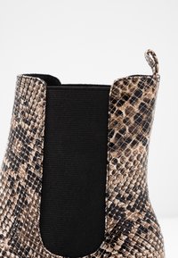 Gros plan sur une botte en cuir imprimé serpent brun et noir avec un panneau latéral élastique noir et une languette de traction sur fond blanc.