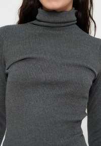 Femme portant un pull à col roulé côtelé gris foncé à manches longues, montrant le haut du torse et la zone du cou.