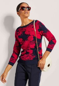 Gebreide marineblauwe trui met een gedurfde rode bloemdesign, ronde halslijn, driekwart mouwen, gecombineerd met een crèmekleurige handtas en oversized rode zonnebril.