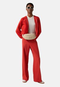 Czerwony komplet z kurtką i spodniami, jasnobeżowa nerka, biały top, okrągłe białe okulary przeciwsłoneczne, płaskie buty; gładka tkanina i dopasowany krój.