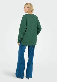 Sweatshirt oversized di un verde scuro con spalle cadenti, orlo a costine e maniche ampie, abbinato a jeans in denim blu a vita alta e a zampa.