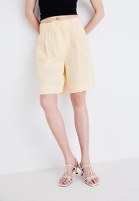 Pantaloni corti giallo chiaro su misura con taglio dritto, davanti plissettato e passanti per cintura, abbinati a sandali bianchi con cinturini su uno sfondo neutro.