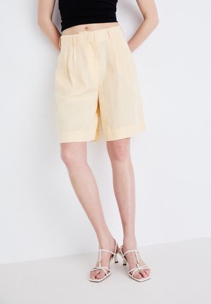Shorts amarillos claros a medida con corte recto, frente con pliegues y presillas para cinturón, combinados con sandalias blancas con tiras sobre un fondo neutro.