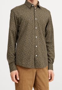 Camicia a maniche lunghe marrone con motivo, arricchita da piccole macchie bianche e verdi, colletto button-down e bottoni bianchi, abbinata a pantaloni in corduroy beige.