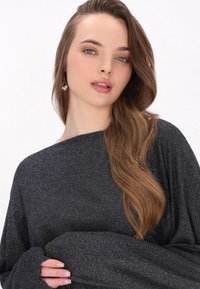 Zwarte, sprankelende oversized top met lange mouwen, bootneck en een zachte textuur. Het haar is gestyled in losse golven. De oorbellen hebben ronde accenten.