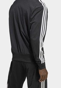 Svart sportjacka med vita tre-rands detaljer längs ärmarna, med en slät textur och figurnära passform.