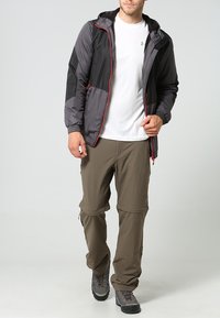 Giacca leggera da uomo nera e grigia con dettagli rossi, abbinata a pantaloni cargo beige e stivali da trekking grigi. Tessuto liscio, design con cappuccio.