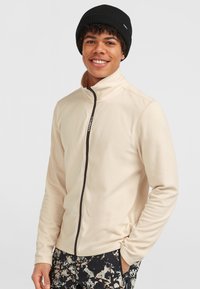 Lys beige fleecejakke med høj krave og brun lynlås; båret med en sort ribbet hue. Mønstrede sort-hvide bukser.