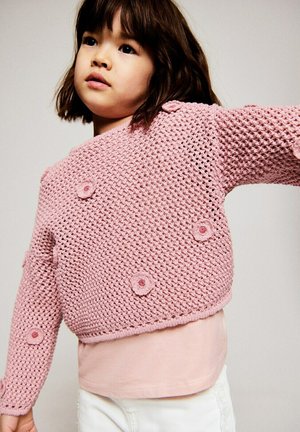 Young girl wearing a pink crocheted sweater with small flower decorations, pale pink shirt, and white pants against a neutral background.