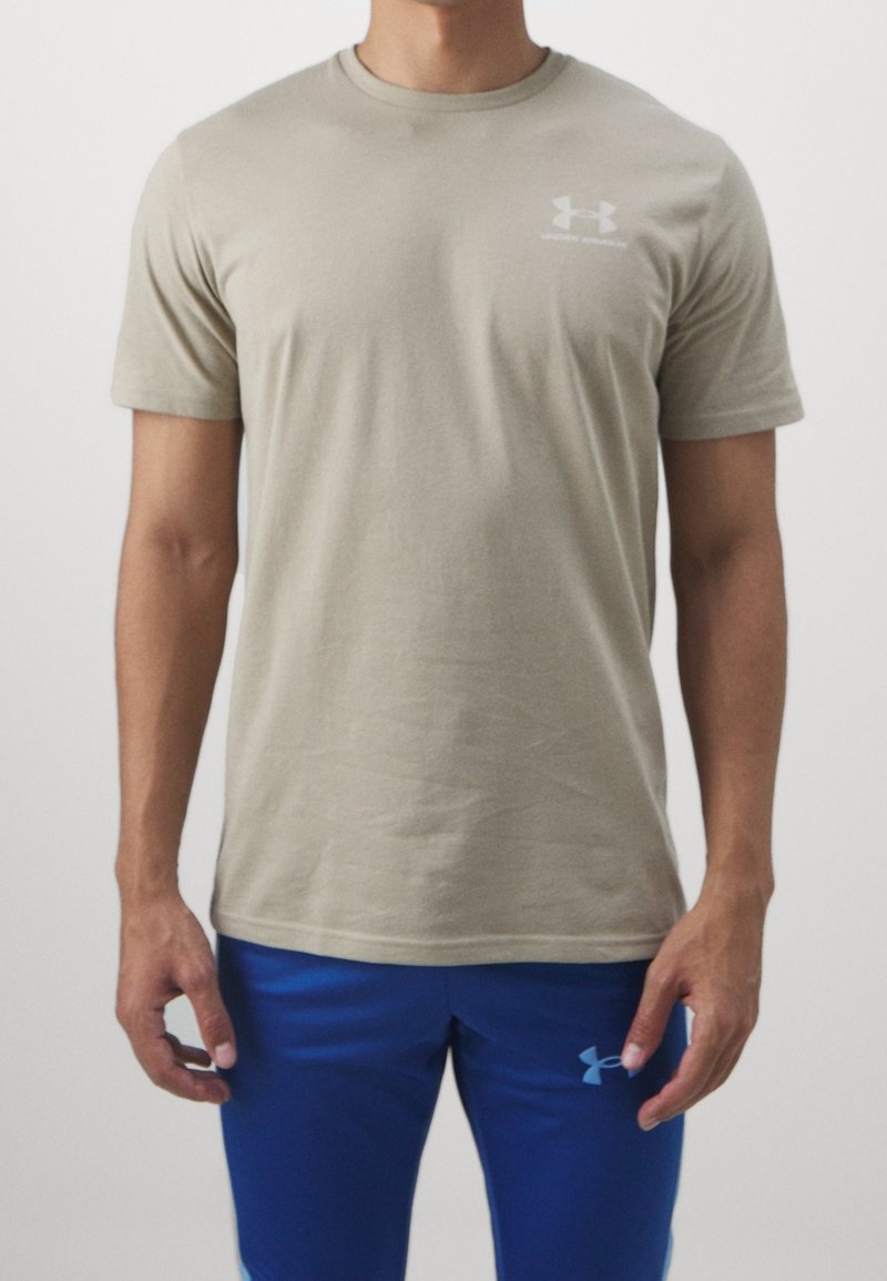 Homme portant un t-shirt manches courtes beige Under Armour et un short de sport bleu Under Armour, debout devant un fond clair uni.