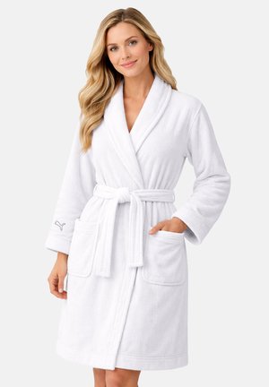 Frau mit langen blonden Haaren, die einen weißen Plüschbademantel mit Taschen und einem gebundenen Gürtel vor einem schlichten hellen Hintergrund trägt.