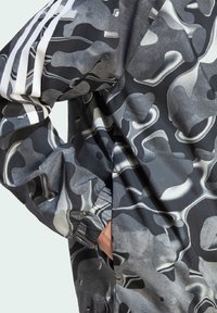 Camouflage-patterned jacket in shades of grey and black, featuring elastic cuffs and three white stripes on the shoulder. Smooth texture.