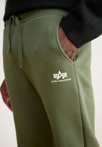 Olivenfarvede sweatpants med snor i taljen, sidelomme og hvidt "ALPHA INDUSTRIES" logo trykt på låret. Blødt bomuldsblandingsmateriale.