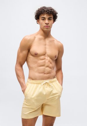 Joven de pie con las manos en los bolsillos, vistiendo un bañador a rayas amarillas, sobre un fondo claro y liso.