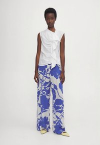 Balts bezroku tops ar volānu detaļām, kombinēts ar plata piegriezuma biksēm ar zilām ziedu rakstiem uz gaiša fona. Gaiši dzelteni apavi.