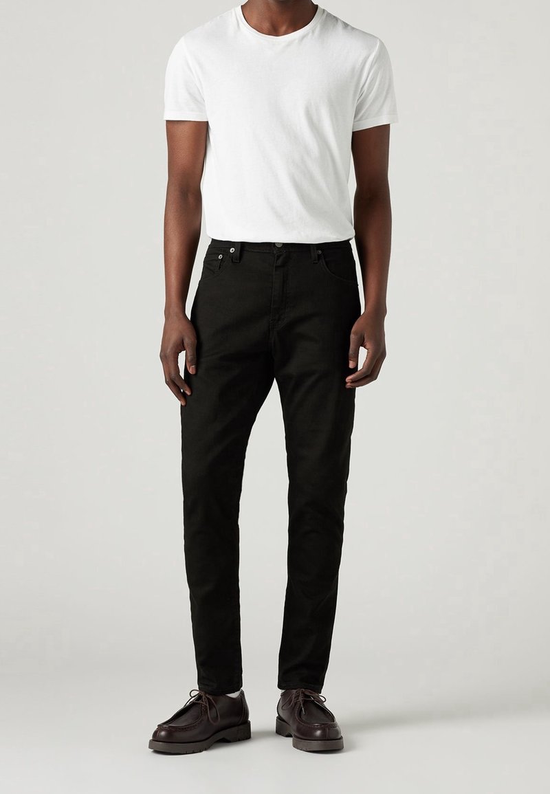 Homme debout portant un t-shirt blanc uni, un jean noir slim-fit et des chaussures lacées marron foncé sur un fond uni.