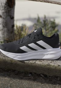 Chaussure de course noire avec des bandes blanches et une semelle blanche épaisse reposant sur une surface en pierre à l'extérieur avec un arrière-plan flou de verdure.