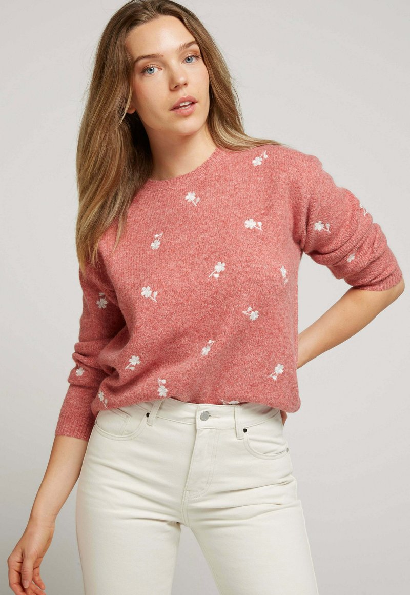 Pull rose clair avec broderies florales blanches, col rond et manches longues, associé à un jean blanc pour un look décontracté.
