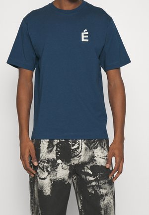 Hombre con camiseta azul marino de manga corta con una pequeña letra blanca acentuada É en el pecho izquierdo y pantalones con estampado en blanco y negro.
