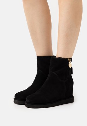 Bottes compensées en daim noir avec fermeture éclair latérale et breloque dorée portées sur des jambes nues sur fond blanc.