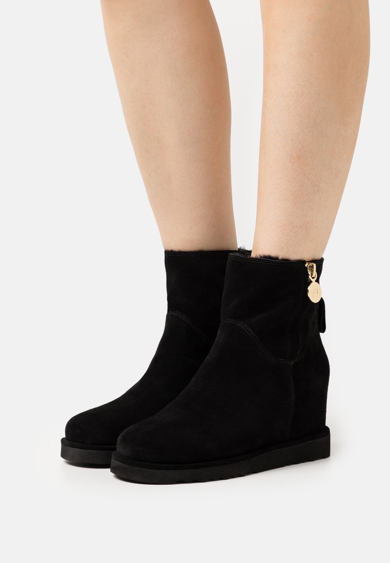 Bottes compensées en daim noir avec fermeture éclair latérale et breloque dorée portées sur des jambes nues sur fond blanc.