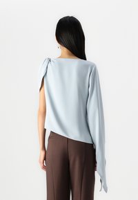 Blusa azzurro chiaro con design drappeggiato, caratterizzata da maniche annodate e taglio dritto, abbinata a pantaloni marroni su misura.