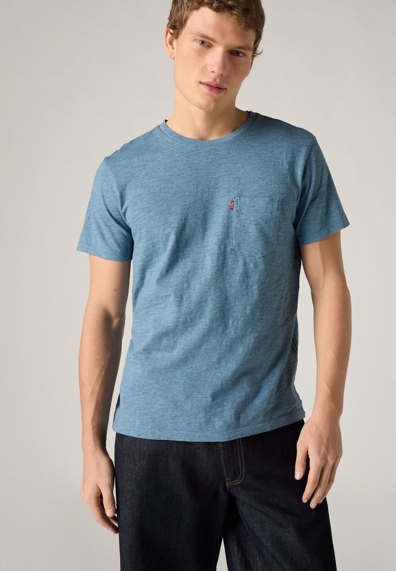 Joven con una camiseta azul de manga corta con bolsillo en el pecho y vaqueros oscuros, de pie frente a un fondo liso.