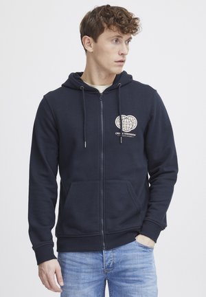 Marineblå hettegenser med glidelås laget av myk tekstil, med to lommer foran og et hvitt globe-design med tekst på brystet. Ribbestrikkede mansjetter.