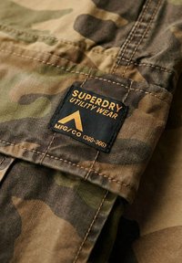 Tela utilitaria de patrón de camuflaje que presenta una etiqueta negra con texto naranja. Costuras visibles a lo largo de los bordes; los materiales parecen duraderos y texturizados.