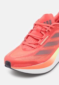 Crvene atletske cipele s mrežastim gornjim dijelom, sa tri tamne pruge. Zaobljen oblik, teksturirana površina i višebojni potplat koji prelazi u crnu boju.