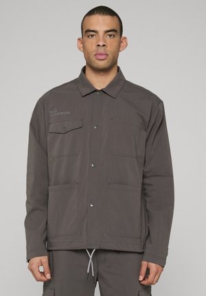 Grå jakke med lange ærmer, knaplukning, to brystlommer og to forlommer. Har logodetalje på venstre side.