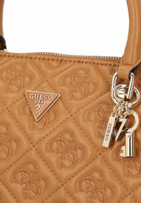 Sac à main matelassé beige avec motif embossé, quincaillerie en ton argent, plaque logo triangulaire, fermeture éclair et accents de porte-clés.