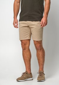 Shorts en coton beige à coupe droite, avec une texture lisse. Associés à des baskets en daim marron et un t-shirt vert foncé.