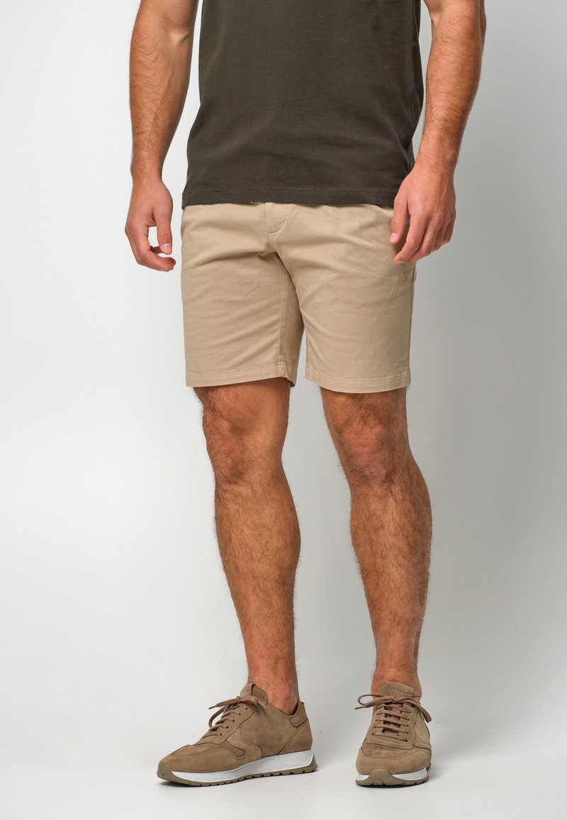 Shorts en coton beige à coupe droite, avec une texture lisse. Associés à des baskets en daim marron et un t-shirt vert foncé.
