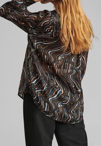 Blusa nera con motivi multicolori a spirale in arancio, blu e bianco. Maniche lunghe con vestibilità ampia, dotata di orlo asimmetrico e texture trasparente.