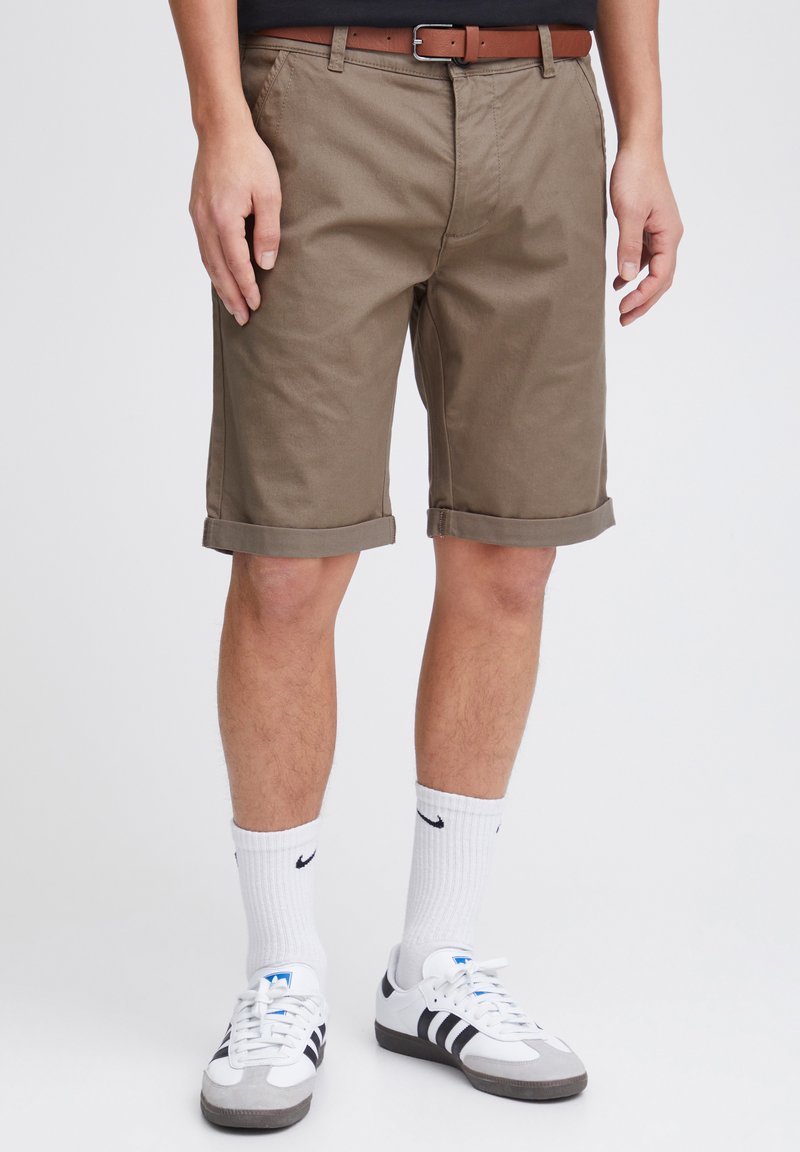 Pantalones cortos de algodón marrón con puños enrollados, bolsillos frontales y un cinturón marrón. Llevados con calcetines blancos y zapatillas en blanco y negro.