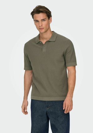 Uomo con polo a maniche corte in maglia verde oliva e jeans blu scuro, in piedi contro uno sfondo chiaro e uniforme.