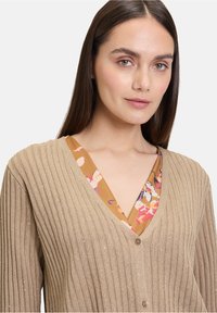Hellbraunes, geripptes Cardigan mit V-Ausschnitt, das einen sanften, metallischen Glanz aufweist und einen bunten Blumenprint auf der Innenschicht hat.