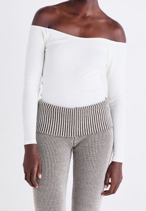Osoba o ciemnej karnacji, ubrana w biały top z długim rękawem odsłaniający ramiona oraz beżowe, prążkowane spodnie z wysokim stanem na jednolitym tle.